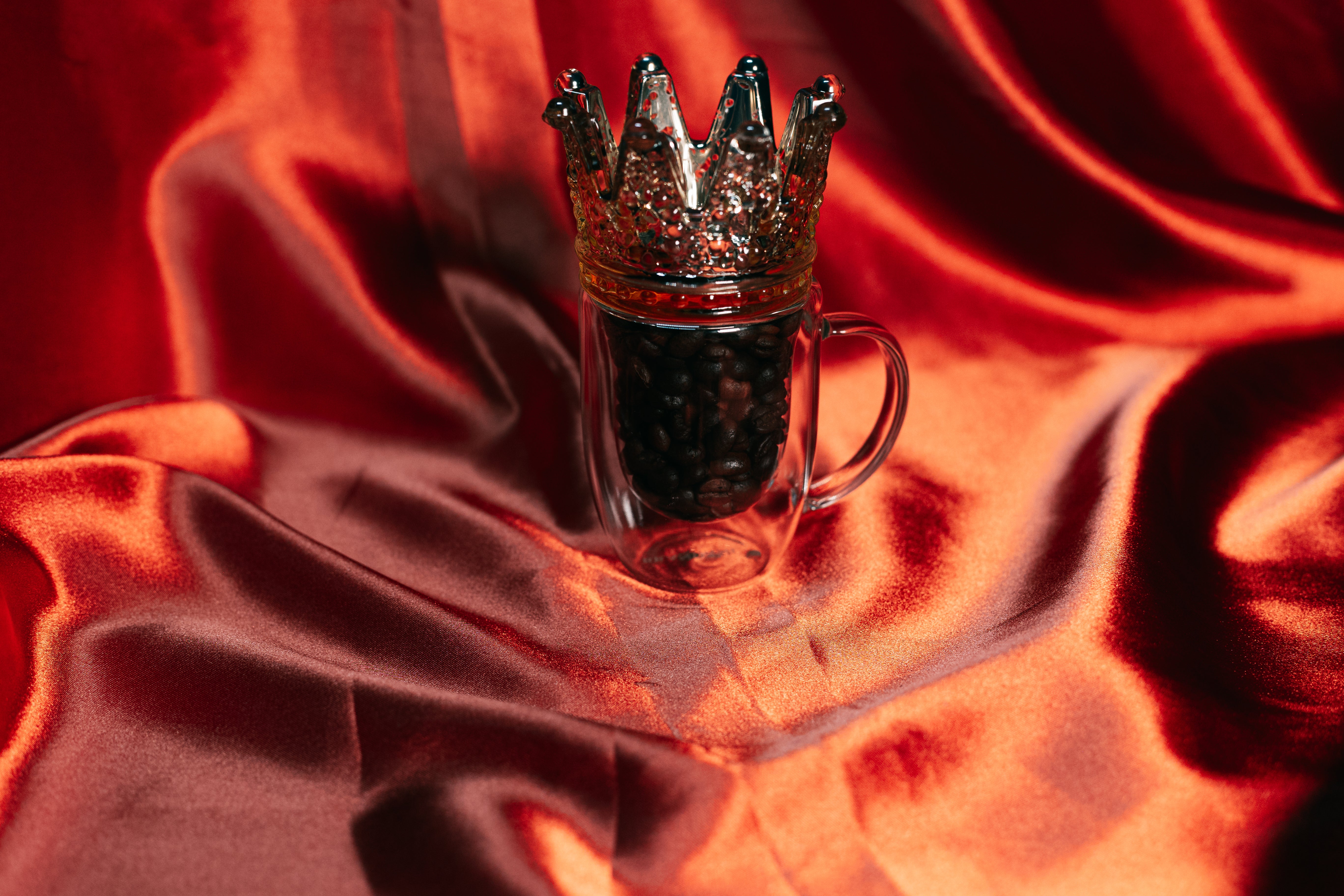 A majestic glass mug filled with Coffee Kingz beans and topped with a king crown, set against a rich red satin backdrop, suggesting royalty or the regal nature of Coffee Kingz to its enthusiasts.